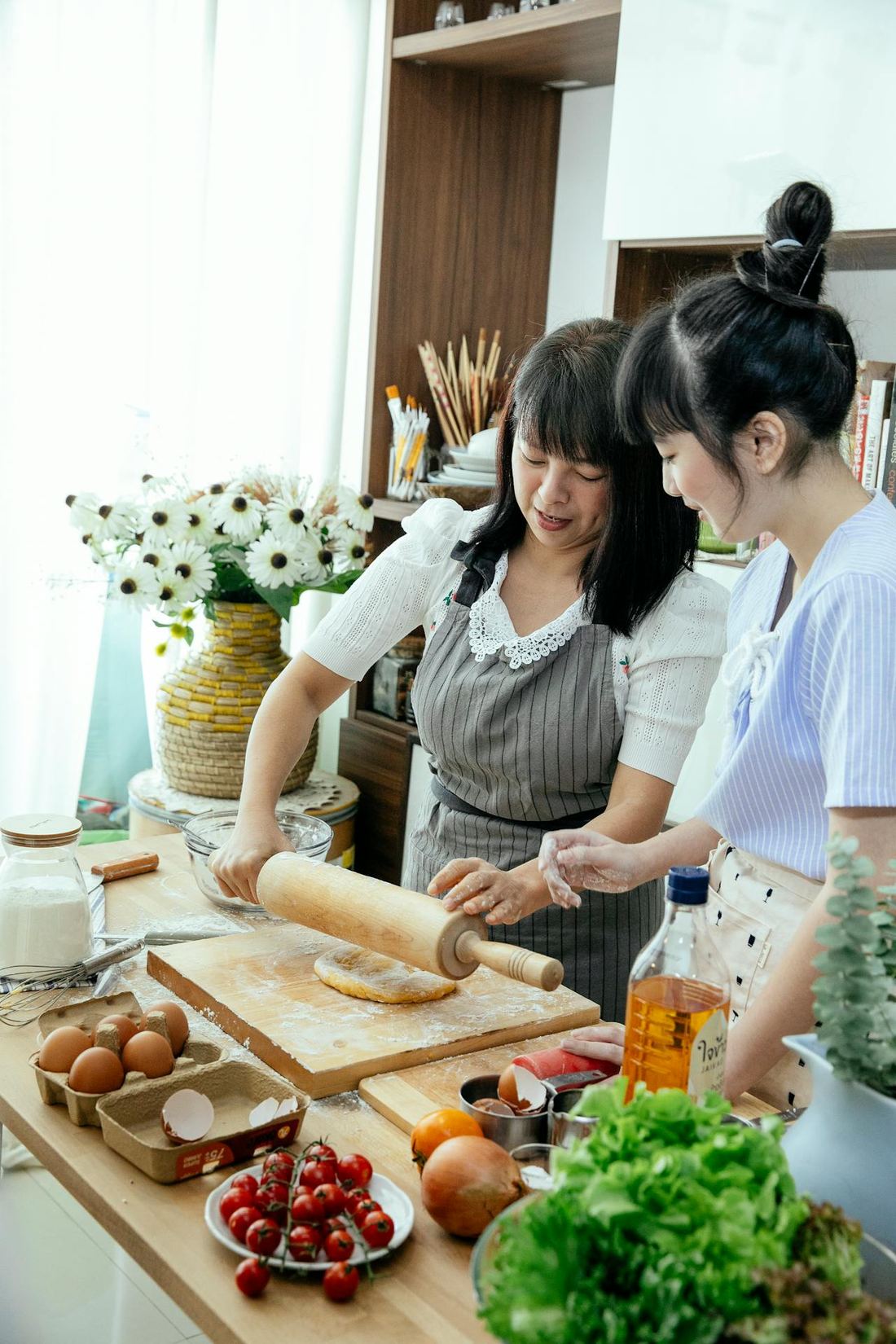 Two women rolling dough together at a kitchen counter, fresh produce around them